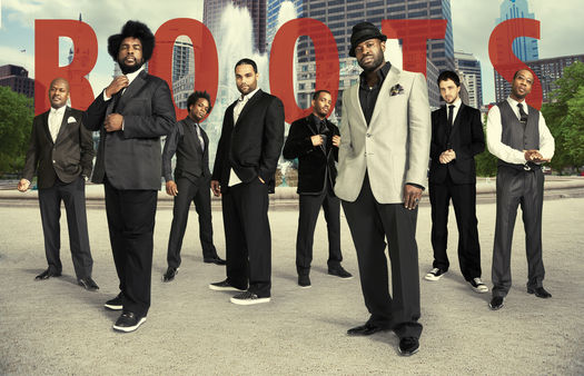 The roots band standing in front of the city skyline with red Roots lettering in the background.